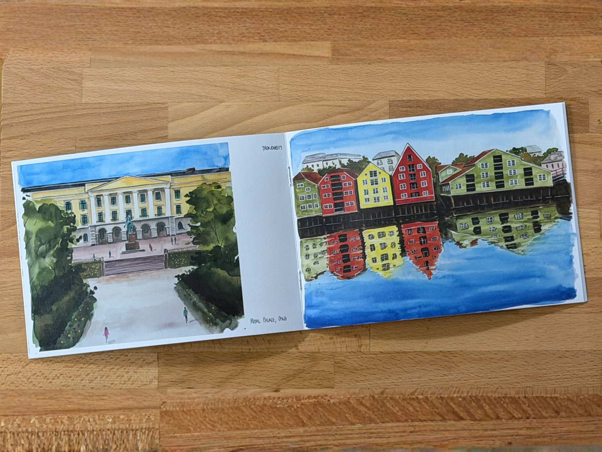 An open watercolor painting book lying on a wooden table, showing two illustrated scenes: a classical building courtyard on the left and colorful waterfront buildings with reflections on the right.