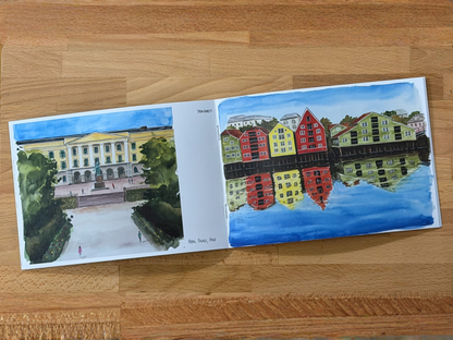 An open watercolor painting book lying on a wooden table, showing two illustrated scenes: a classical building courtyard on the left and colorful waterfront buildings with reflections on the right.