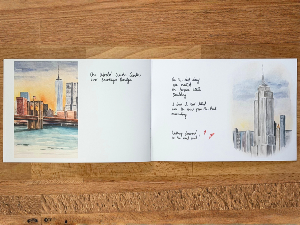 An open watercolor sketchbook lying flat on a wooden surface, showing two illustrated city skyline pages with handwritten notes in the center.