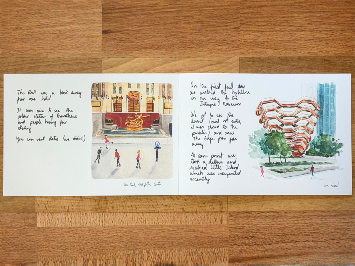A printed art card or folded sheet with handwritten and illustrated notes about The Rock in NYC, placed on a wooden tabletop. The sheet includes a small cityscape drawing, a red-and-gold fountain illustration, and a watercolor-style depiction of the Rock with surrounding trees and buildings.
